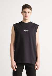 Juoda be rankovių marškinėliai, pagaminti iš medvilnės. Turi apvalų kaklą ir mažą baltą logotipą su užrašu "Calvin Klein Jeans" ant krūtinės.