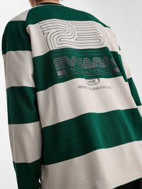 Osoba ubrana w zielono-biały szeroko paskowany długi rękaw z napisem "23 SWARM INTERNATIONALS" i "Serving the game since 1923" na plecach.