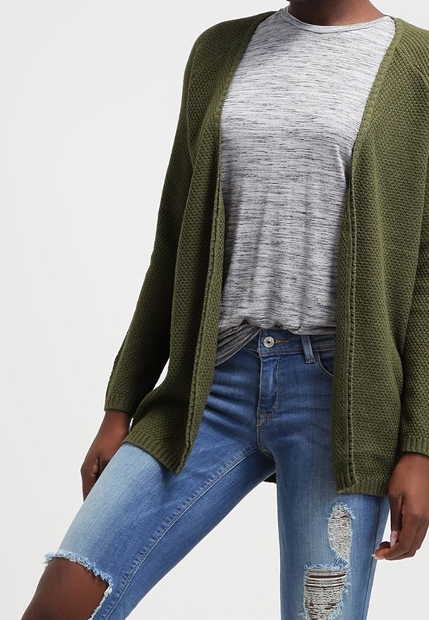 Oliivinvihreä neulecardigan avonaisella edellä, jossa on teksturoitu kuvio. Musta harmaanraidallisen paidan päällä, yhdistettynä kulutettuihin sinisiin farkkuihin.