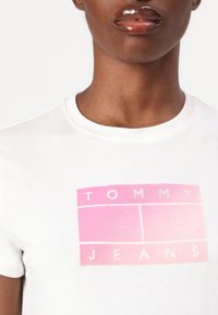 T-shirt en coton blanc avec un logo dégradé rose et le texte "TOMMY JEANS" ; col rond, manches courtes, texture lisse.
