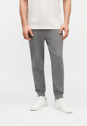 Mees, kes kannab halli jooksupükse, valgeid tossusid ja valget T-särki, seisab ühevärvilise heleda tausta ees.