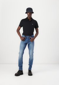 Tricou polo negru, blugi decolorat albastru deschis și cizme negre masive. Modelul poartă ochelar de soare întunecați. Outfit simplu cu accente minime.