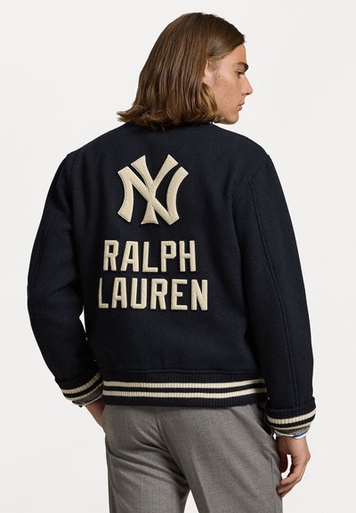 Marineblå uld varsityjakke med beige NY-logo og "RALPH LAUREN" broderet på ryggen, ribbede manchetter og kant med stribede detaljer.