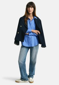 Marineblauwe dubbelbettjacket met gouden knopen, lichtblauwe button-up shirt en vervaagde rechte jeans, gecombineerd met witte sneakers.