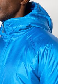 Modra kapuca iz lahkega, sijajnega materiala. Ima visok ovratnik, sprednji zadrgo in logotip blagovne znamke na rami.