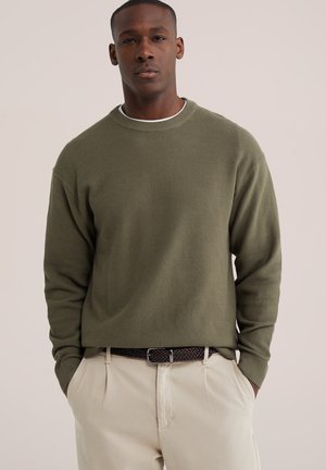 Mand med kort hår iført en olivengrøn tekstureret sweater, beige bukser og et mørkt flettet bælte, stående med hænderne i lommerne.