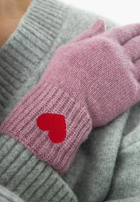 Roze gebreide handschoen met geribde manchet en een rode hartpatch, geplaatst tegen een zachte grijze trui. Gecodeerd breipatroon.
