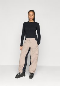 Pantaloni de schi bej și negri, cu fermoare și buzunare, asortați cu un top negru cu mânecă lungă, croită pe corp. Modelul stă în picioare, purtând cizme de schi.