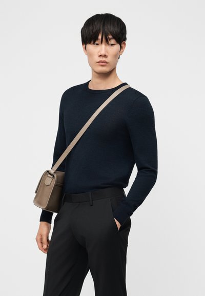 Man met een zwart shirt met lange mouwen en broek, met een beige crossbodytas, kijkend naar de zijkant tegen een effen achtergrond.