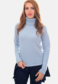 Femme aux longs cheveux auburn vêtue d'un pull col roulé bleu clair et d'une jupe plissée bleu marine se tenant devant un fond blanc.