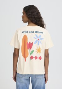 Bež bombažna majica s pisanim grafičnim potiskom cvetlice, lista, sonca in geometrijskih oblik na hrbtni strani ter napisom "Wild and Bloom".
