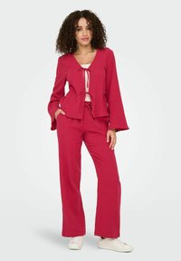 Completo due pezzi di lampone con top a maniche a campana e nodo frontale, abbinato a pantaloni a gamba larga, realizzati in tessuto texturizzato. Sneakers bianche.