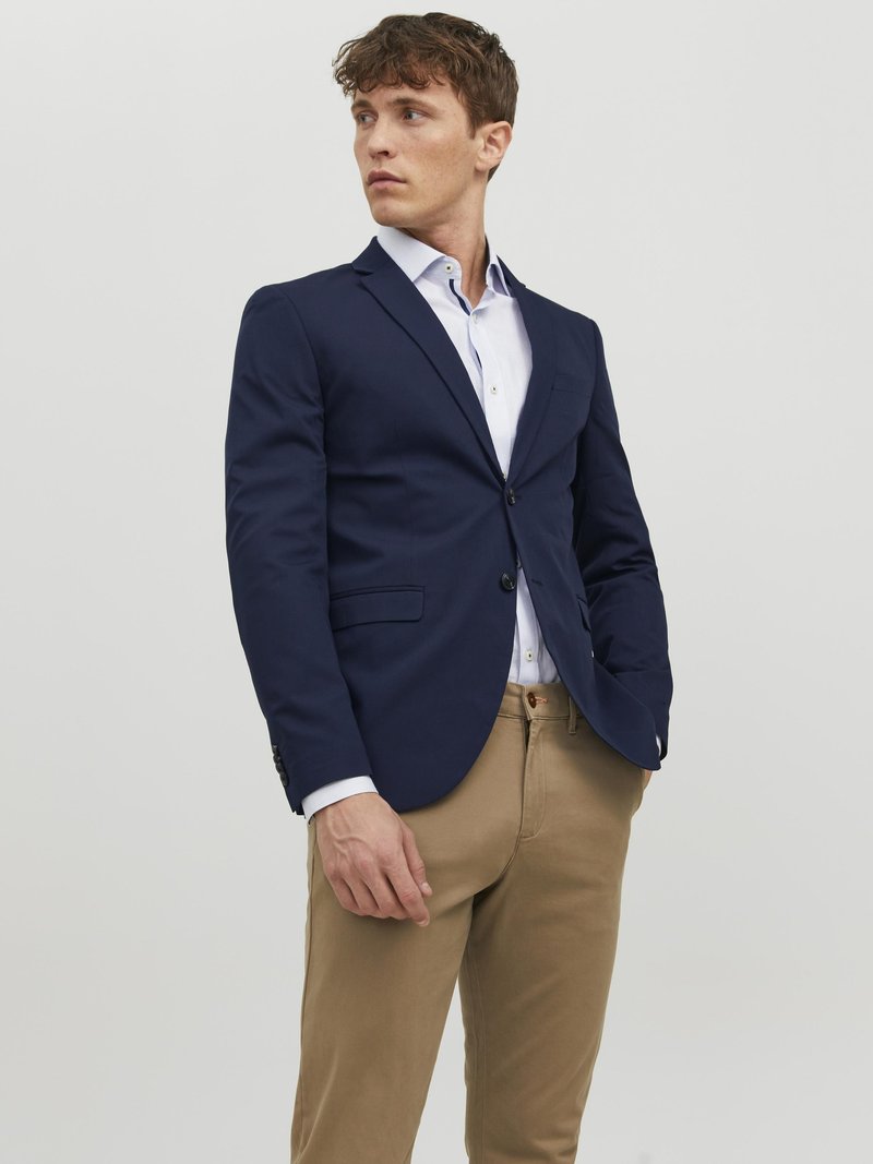 Blazer bleu marine avec fermeture à un bouton, détails de revers, porté sur une chemise à boutons claire et un pantalon beige. Tissu lisse avec coupe ajustée.
