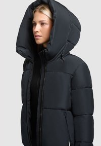 Puffjacka i mörk marinblå färg, med en stor huva, quiltad design, dragkedjestängning och sidofickor. Mjukt, slätt tyg med en elegant silhuett.