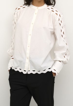 Personne portant un chemisier blanc boutonné avec des motifs découpés sur les manches et un ourlet festonné, associé à un pantalon noir.