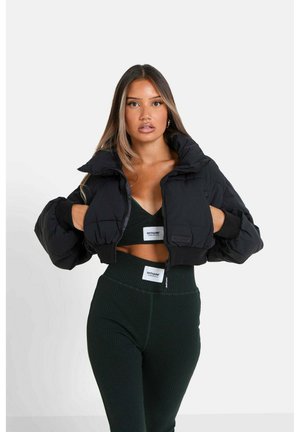 Veste courte matelassée noire avec un col haut et des poignets côtelés, dotée d'une fermeture éclair à l'avant et d'un patch de marque sur la manche. Portée sur une tenue côtelée vert foncé.
