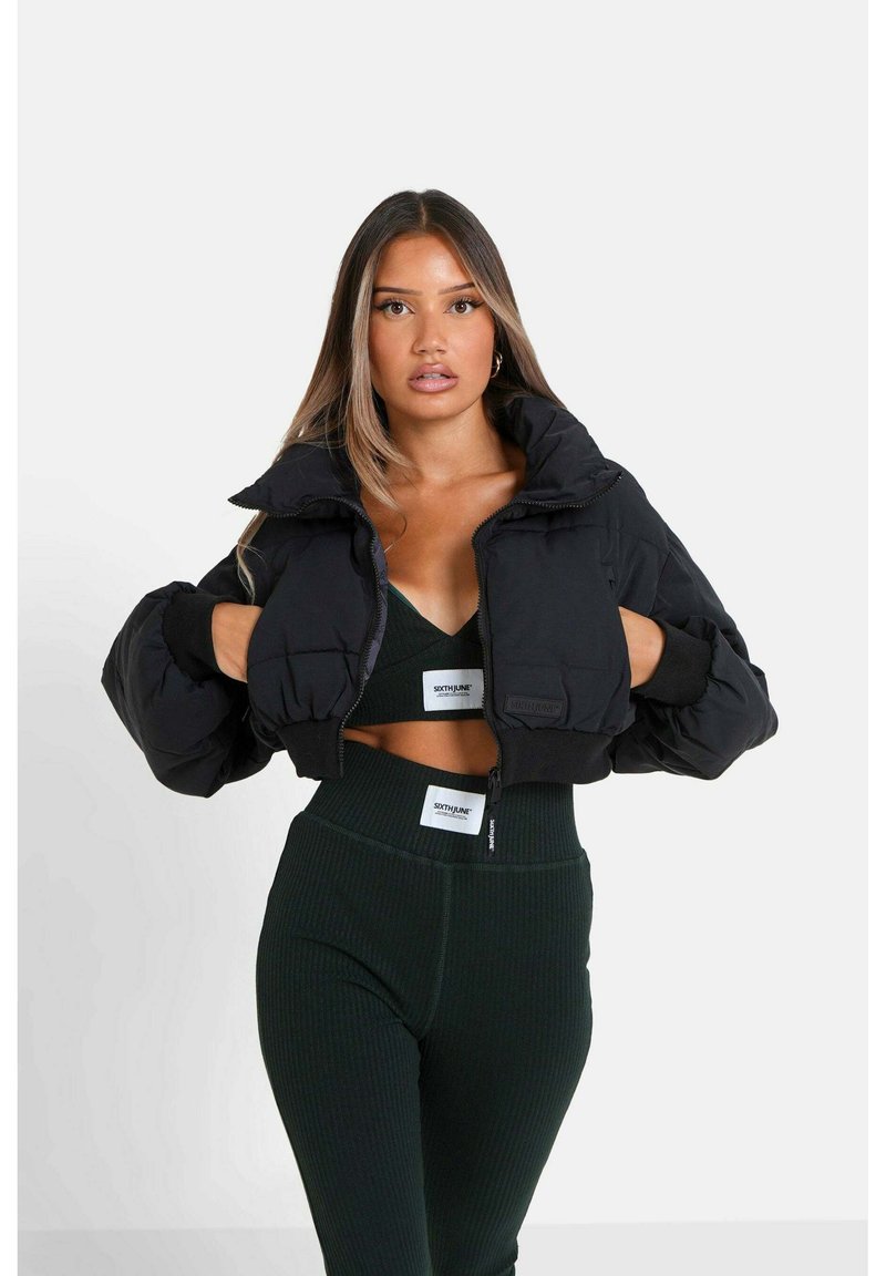 Chaqueta corta de plumón negra con cuello alto y puños de canalé, con cremallera frontal y parche de marca en la manga. Lleva puesta sobre un conjunto de canalé verde oscuro.