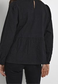 Černý bavlněný top s dlouhým rukávem a peplum pasem, texturovaná tkanina a detail klíčové dírky na zádech. Upnuté rukávy a základní struktura.