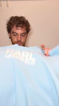 Άνδρας με σγουρά μαλλιά κρατά ένα ανοιχτό μπλε πουκάμισο με μερικώς ορατό λευκό κείμενο σε απλό μπεζ φόντο.