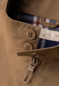 Col de veste beige avec deux boutons et tirette de fermeture éclair, tous de marque "OMBRE", montrant une doublure à carreaux bleus et marron à l'intérieur.