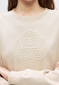 Sweat-shirt beige avec un motif géométrique circulaire texturé sur la poitrine, porté par une personne aux longs cheveux bruns et les bras croisés.
