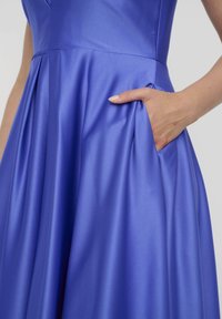 Ein lebhaftes, blaues Kleid mit einer glatten, satinierten Textur, einem taillierten Oberteil, einem fließenden Rock und einer Hand in einer Seitentasche.