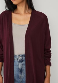 Bordeaux open-front cardigan gemaakt van een zachte breisel, met geribde manchetten en een comfortabele pasvorm, gedragen over een licht tanktop en blauwe jeans.