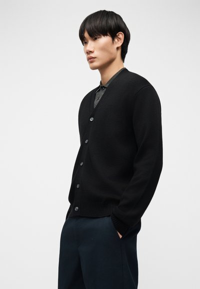 Νέος άνδρας που φορά μαύρο ζακέτα με κουμπιά πάνω από σκούρο γκρι πουκάμισο, με τα χέρια στις τσέπες, στέκεται μπροστά σε απλό λευκό φόντο.