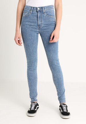Personne portant un jean skinny taille haute bleu clair et des baskets noires et blanches, debout les mains détendues le long du corps.