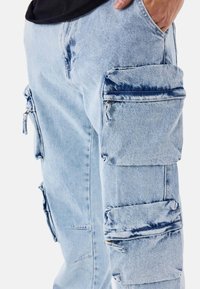 Pantalones cargo de mezclilla azul claro con múltiples bolsillos con cremallera y detalle de correa ajustable en el lateral. Textura desgastada en la tela.