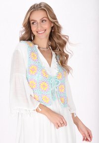 Häkelweste mit einer pastellfarbenen Palette in Rosa, Gelb und Blau mit floralen Mustern, vorne gebunden. Über einem weißen Kleid getragen.