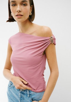 Kvinde iført en ærmeløs lyserød top med en guldfarvet spænde på den ene skulder, kombineret med lyseblå jeans, stående mod en ensfarvet baggrund.