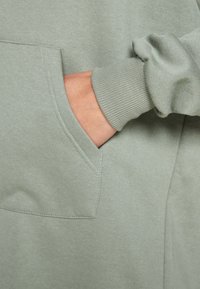 Bavlněná mikina v jemně šalvějové zelené barvě, s přední kapsou, žebrovanými manžetami a hladkým povrchem bez viditelných švů nebo log.