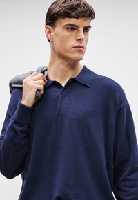 Σακάκι πλέ knit σε ναυτικό μπλε χρώμα με κλασικό γιακά, δύο κουμπιά και μακριά μανίκια, με ομαλή υφή και φθαρμένο γκρι πουλόβερ ριγμένο στον ώμο.