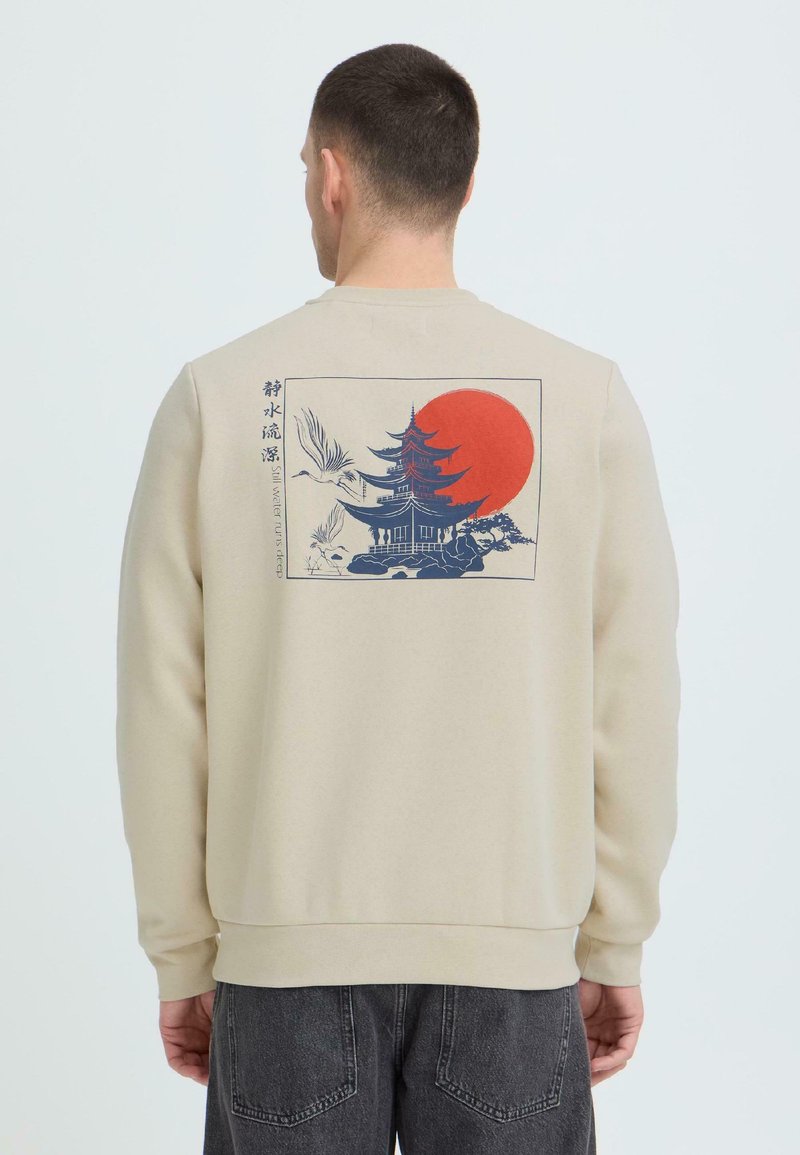 Homme portant un sweat-shirt beige orné d'un motif de pagode, de grues en vol et d'un soleil rouge avec le texte vertical « Still water runs deep » au dos.