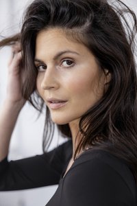 Portret osobe s dugom, tamnosmeđom kosom, koja nosi crnu majicu. Osobine uključuju srednju boju kože i neutralnu šminku.