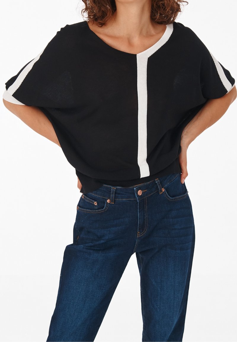 Femme portant un haut noir à manches courtes avec des rayures blanches verticales et sur les manches, et un jean bleu foncé, mains posées sur les hanches.