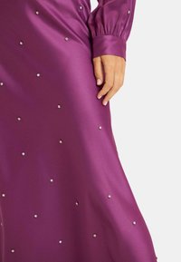 Vestido de satén morado con mangas largas, adornado con tachuelas plateadas dispersas que crean una apariencia texturizada. Acabado suave y brillante.