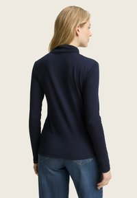 Femme aux cheveux blonds portant un haut à col roulé bleu marine à manches longues et un jean, de dos contre un fond uni.
