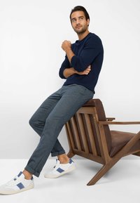 Donkerblauwe langemouwen shirt, donker geruite broek, witte sneakers met blauwe accenten, zittend op een houten stoel met een bruine zitting.