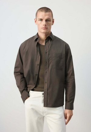 Joven con el pelo corto que lleva una camisa marrón oscuro abotonada sobre una camiseta marrón y pantalones blancos, de pie frente a un fondo claro y liso.