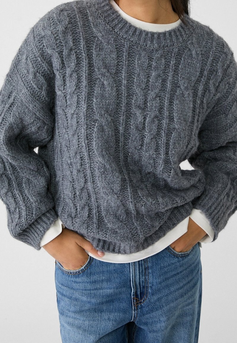 Grå kabelstickad tröja med rund hals och oversize-passform, lagd över en vit skjorta. Har texturerade mönster och ribbade muddar.