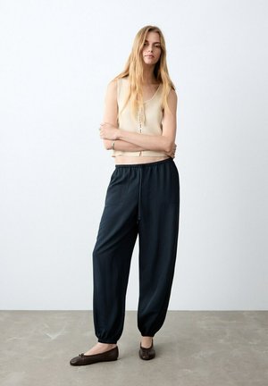 Mujer joven con cabello largo y rubio, vestida con una blusa sin mangas beige, pantalones negros holgados y bailarinas marrones, de pie con los brazos cruzados.