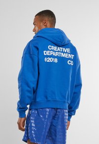 Helle Baumwollkapuzenjacke mit großem weißen Text auf dem Rücken, der "KREATIVABTEILUNG 2018 CD" liest. Enthält eine Kapuze mit Kordelzug und gerippten Saum.