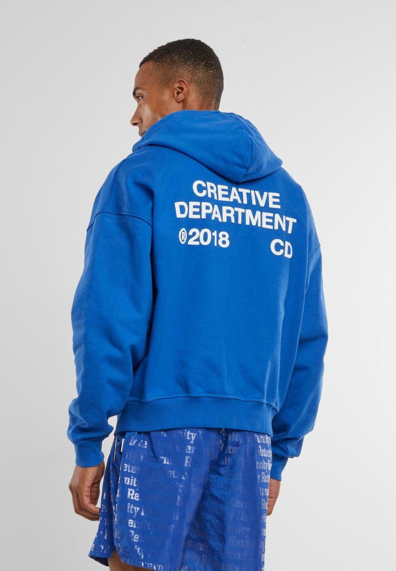Helle Baumwollkapuzenjacke mit großem weißen Text auf dem Rücken, der "KREATIVABTEILUNG 2018 CD" liest. Enthält eine Kapuze mit Kordelzug und gerippten Saum.
