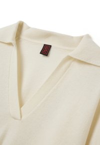 Pull en tricot de couleur crème avec un col en V. Dispose d'un tissu texturé et d'une étiquette noire avec du texte rouge près du col.