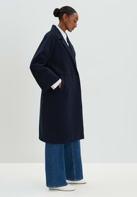 Marineblauer, doppelt geknöpfter Mantel mit weiten Ärmeln, zwei seitlichen Taschen und lockerer Passform, kombiniert mit hellblauen Schlagjeans und weißen Schuhen.