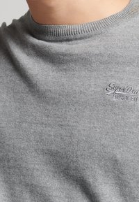 T-shirt en coton gris avec un col côtelé et une texture subtile. Présente un petit logo "SuperDry" brodé dans le coin supérieur gauche.