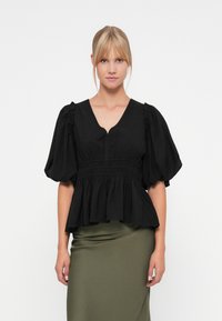 Svart topp med puffermer, V-hals og smocket midje. Glatt bomullsstoff med en flytende peplum-silhuett. Kombinert med en olivengrønn skjørt.