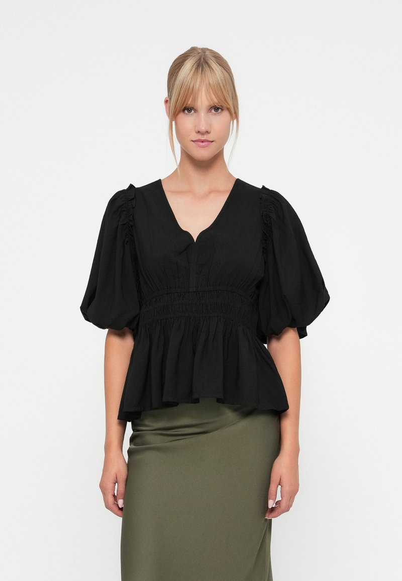 Svart topp med puffermer, V-hals og smocket midje. Glatt bomullsstoff med en flytende peplum-silhuett. Kombinert med en olivengrønn skjørt.